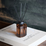 Libra Interiors amber glass 200ml reed diffuser with softly triangular brown vessel, rounded black collar and slim black fibre reeds, set on a style book.