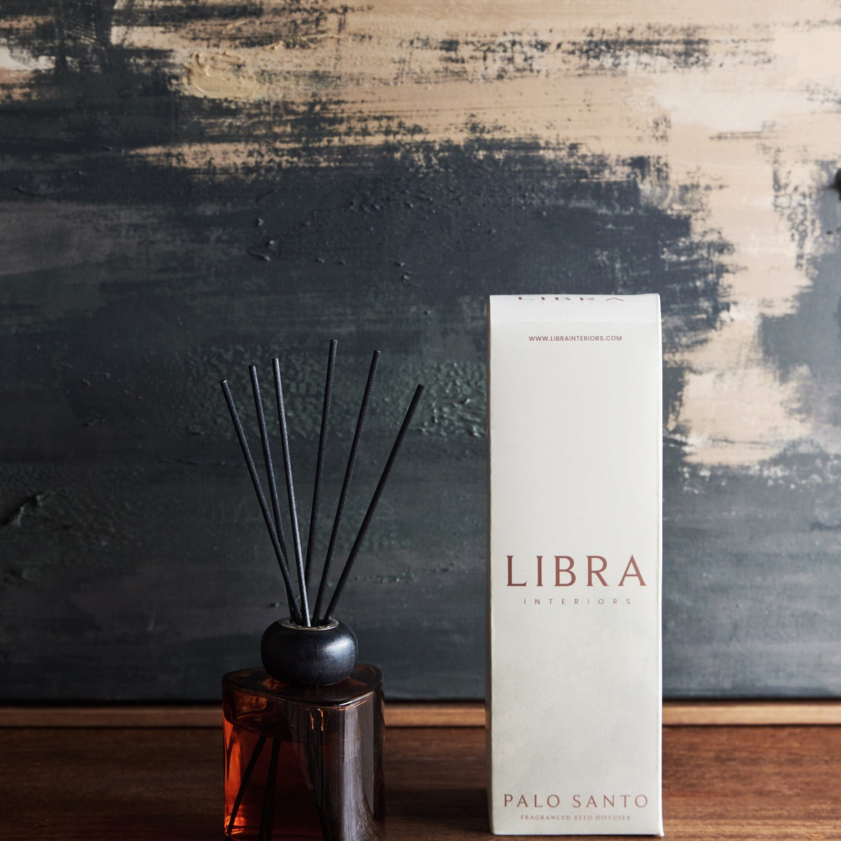 Libra Interiors Palo Santo reed diffuser with amber glass bottle, black collar and fibre reeds, shown beside its white box on a wooden surface.