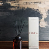 Libra Interiors Palo Santo reed diffuser with amber glass bottle, black collar and fibre reeds, shown beside its white box on a wooden surface.
