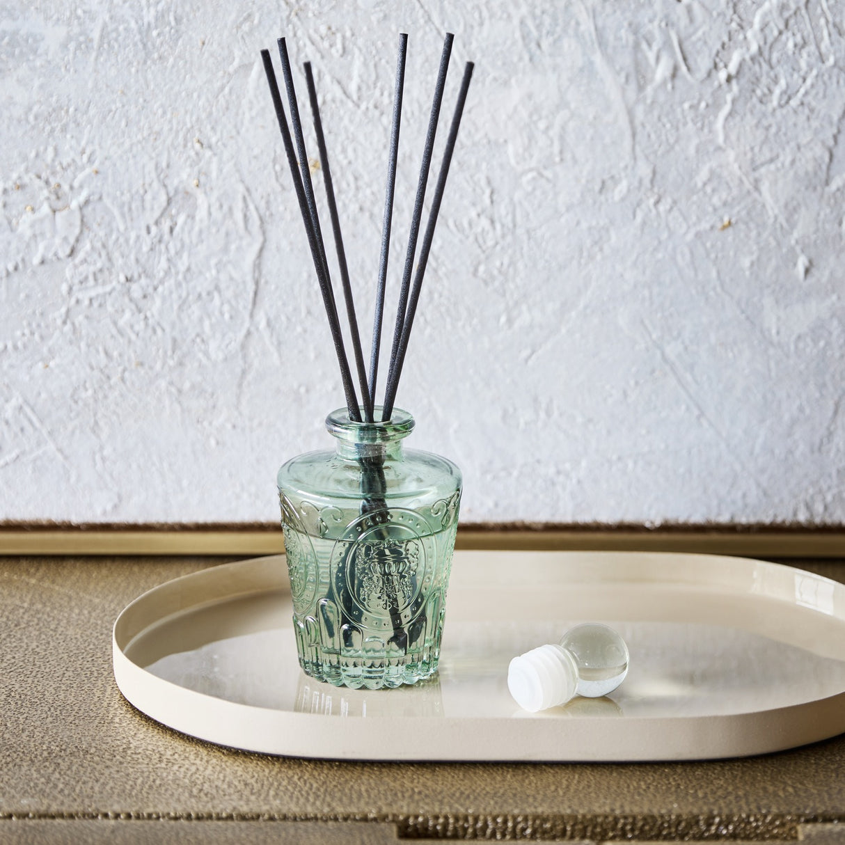 Libra Interiors Embossed Glass Reed Diffuser in soft green tinted glass with black reeds and spherical stopper, set on a cream oval tray.