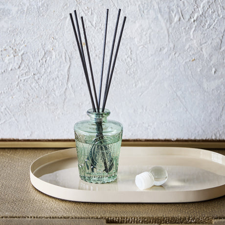Libra Interiors Embossed Glass Reed Diffuser in soft green tinted glass with black reeds and spherical stopper, set on a cream oval tray.