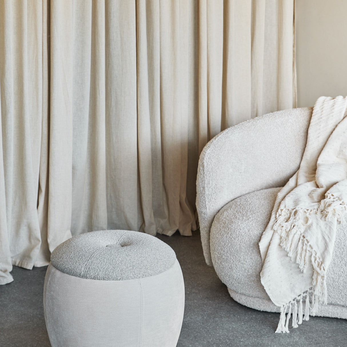 Libra Interiors Velvet Boucle Round Storage Stool in cream beside a boucle armchair with cream throw, set against soft linen curtains in a living room.