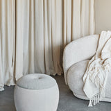 Libra Interiors Velvet Boucle Round Storage Stool in cream beside a boucle armchair with cream throw, set against soft linen curtains in a living room.