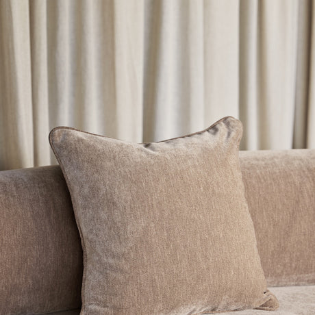 Libra Interiors Piped Plush Velvet Cushion in brown resting on a mocha sofa in front of soft cream curtains, showing corded piping along the seams.