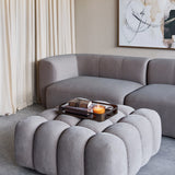 Libra Interiors Velvet Channelled Square Ottoman in cream with a tray, books and lit candle on top, set beside a grey modular sofa in a living room.