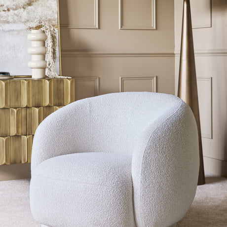 Libra Interiors Bouclé Swivel Accent Armchair in cream with a rounded silhouette, set beside a fluted brass cabinet and tall metallic cone decor.