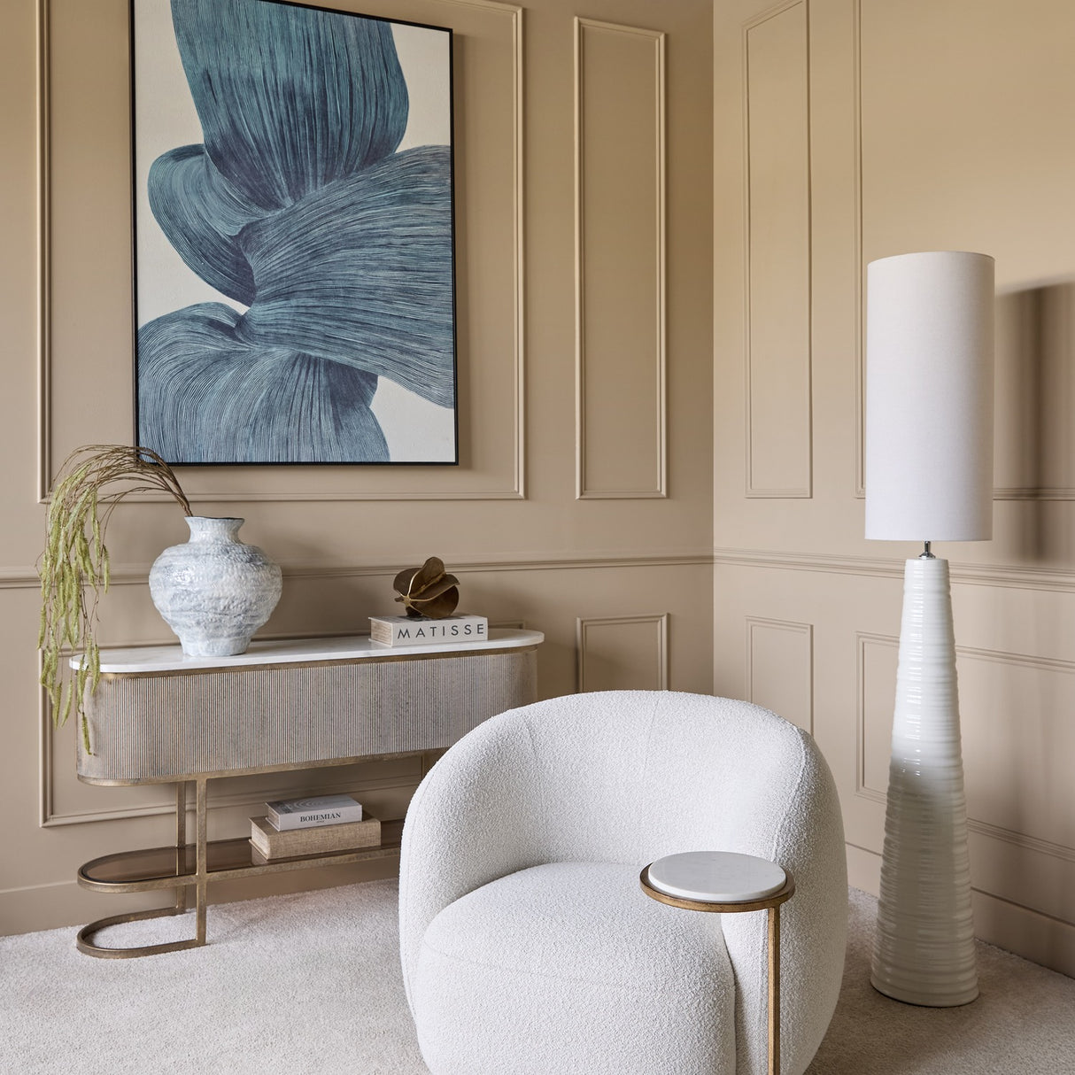 Libra Interiors Bouclé Swivel Accent Armchair in cream beside a fluted console, floor lamp and blue abstract artwork in a panelled beige room.