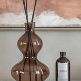 Libra Interiors Palo Santo 500ml reed diffuser refill in an amber-brown bottle with black cap, beside a brown glass diffuser vessel and abstract artwork.