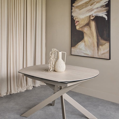 Libra Interiors extendable ceramic dining table in warm stone with brushed grey angular metal legs, styled with a cream vase in a neutral dining setting.