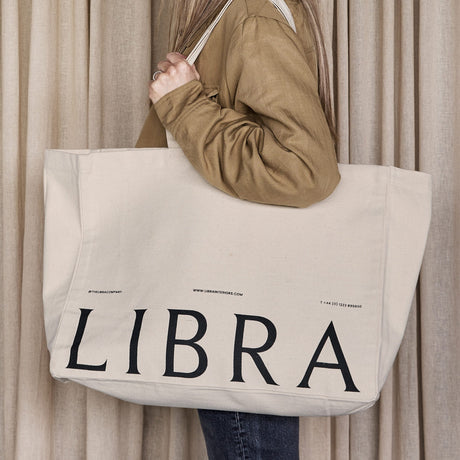 Woman in tan jacket carries Libra Interiors natural cotton tote bag over her shoulder, printed with LIBRA in black lettering, against a beige curtain backdrop.