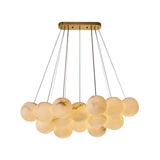 The Noctis 12 Light Medium Floating Globes Alabaster Chandelier in Brushed Brass features a rectangular gold ceiling mount and multiple alabaster globes at varying heights, emitting a warm, soft glow.