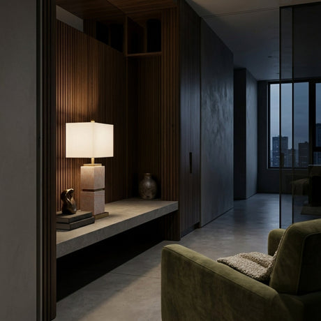 A modern, dimly lit hallway with wood paneling features a concrete bench holding the Veradex 1 Light Marble Block Table Lamp in Brass & Purple, books, a small sculpture, and a vase. An olive green armchair and large city-view windows are in the background.