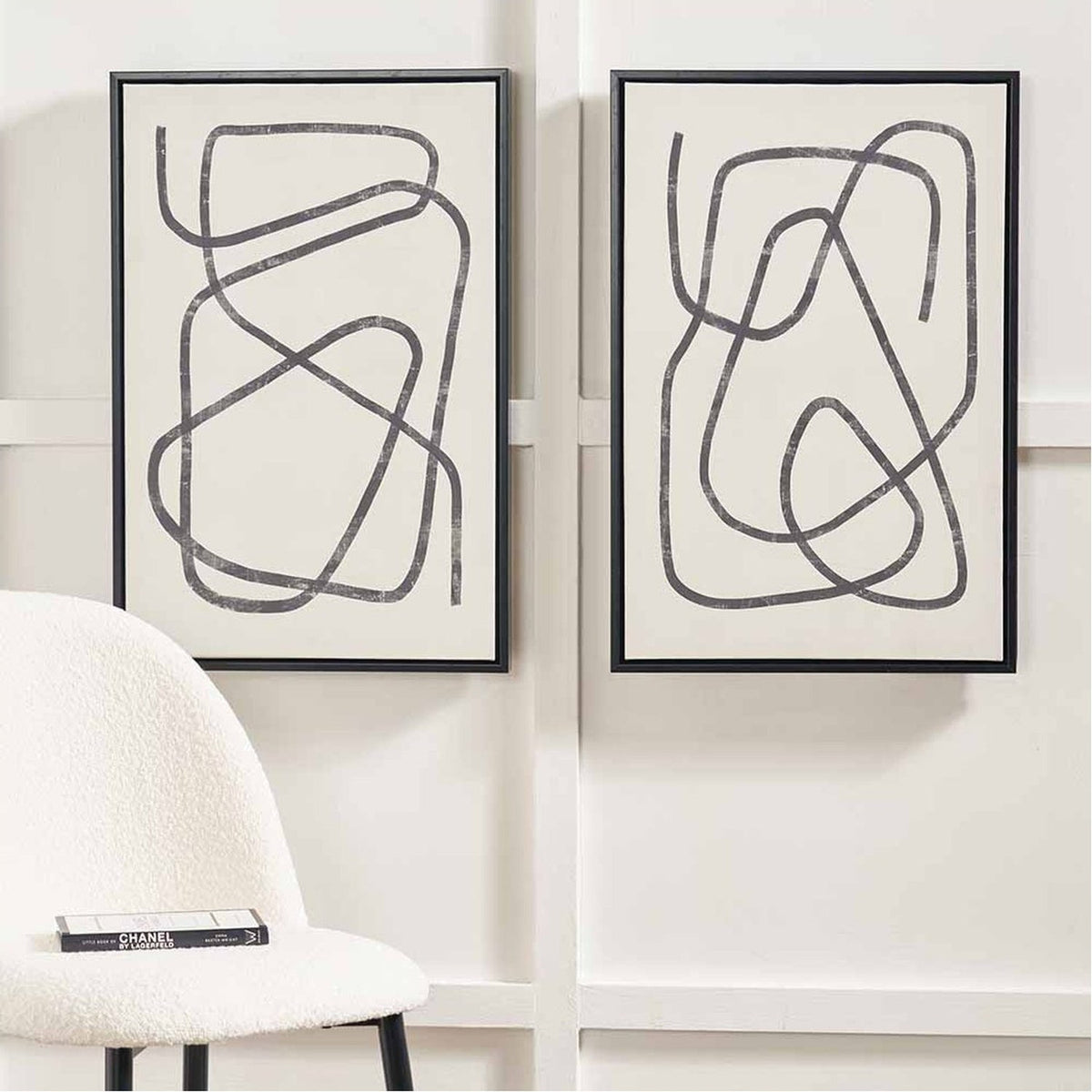 Set of 2 black-framed canvas wall art prints featuring grey squiggle line designs on cream backgrounds, shown mounted on a wall above a boucle armchair.