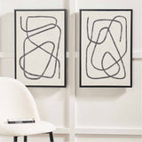 Set of 2 black-framed canvas wall art prints featuring grey squiggle line designs on cream backgrounds, shown mounted on a wall above a boucle armchair.