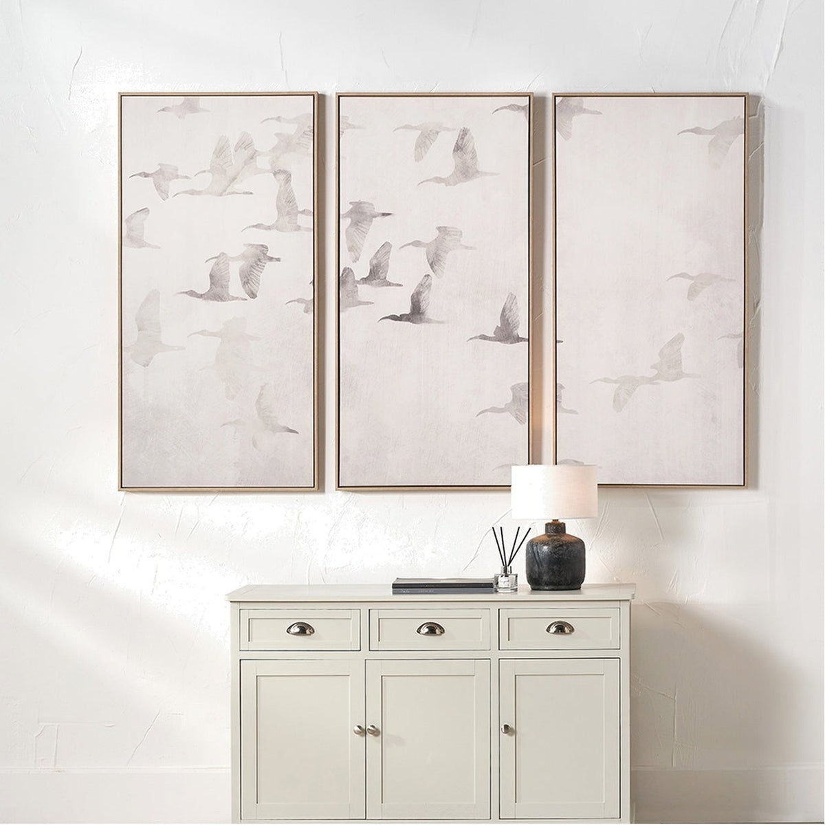 Set of 3 framed canvas wall art panels with flying bird motifs in natural and white tones, hung above a white cabinet in a neutral-toned room.