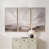 Set of three grey and neutral abstract landscape canvas wall art panels above a light cabinet, each canvas showing soft cloud-like tones over a darker horizon band.