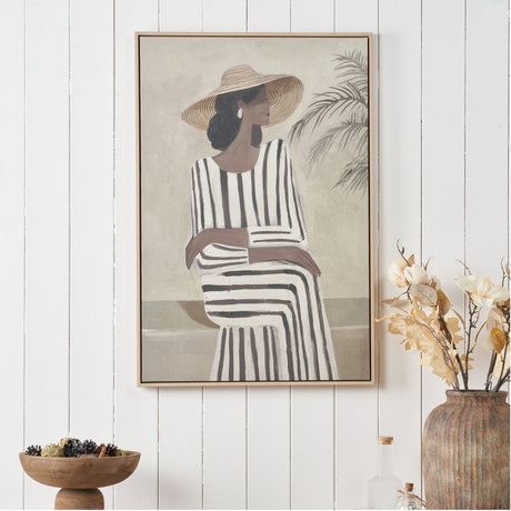 Rectangular canvas wall art in a natural wood frame, depicting a woman in a striped dress and sun hat beside a palm plant, shown hanging on a white panelled wall.