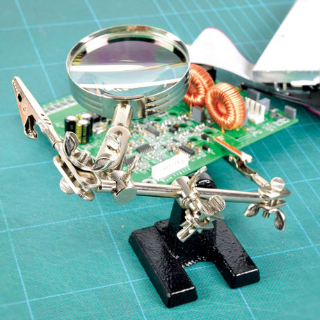 AVSL Mercury MAG01 Helping Hands Magnifier Stand with metal base, two crocodile clips and 60mm glass lens, holding a circuit board on a green mat.