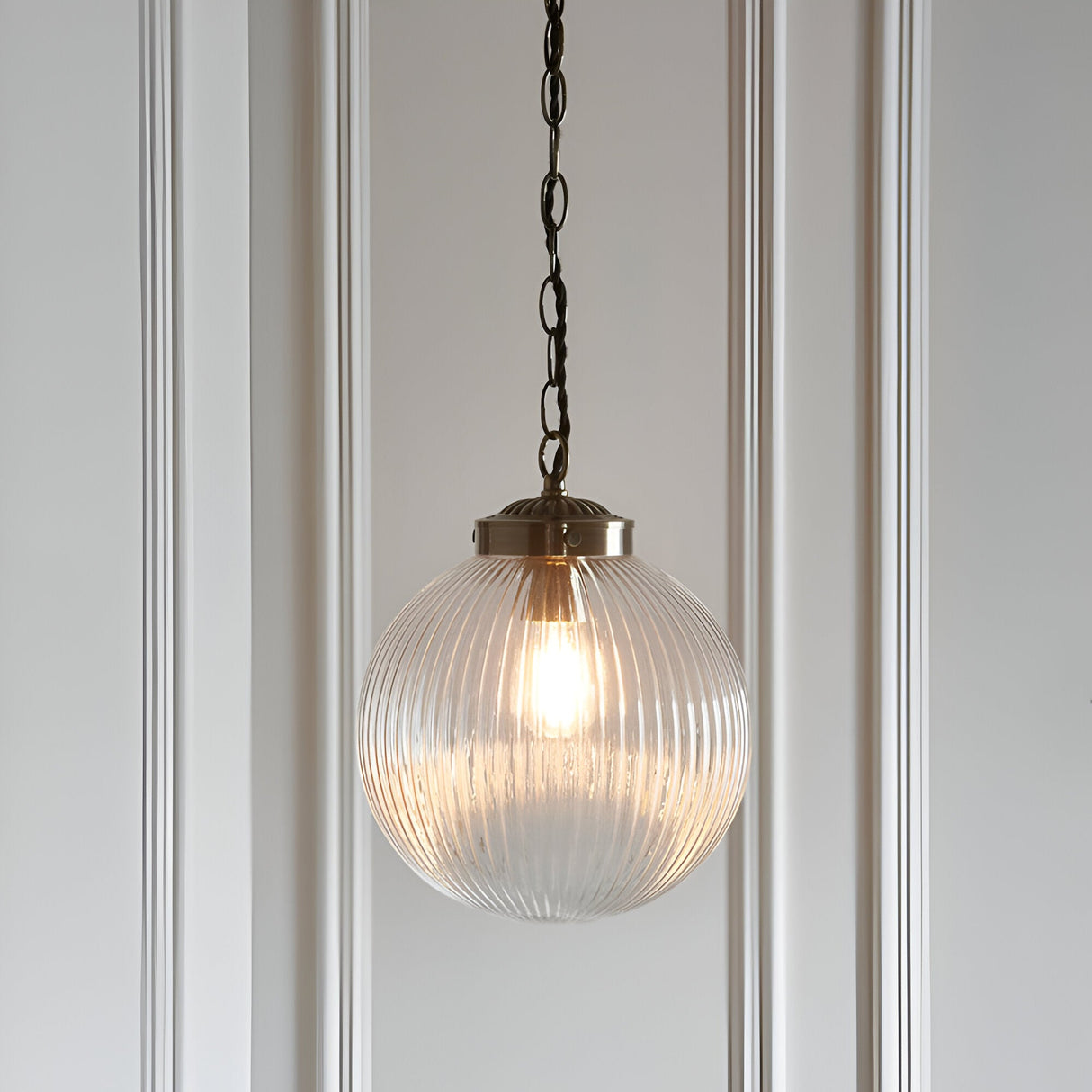 Endon Ashmere pendant light with ribbed clear glass globe shade and antique brass chain suspension, hung against a white panelled wall.