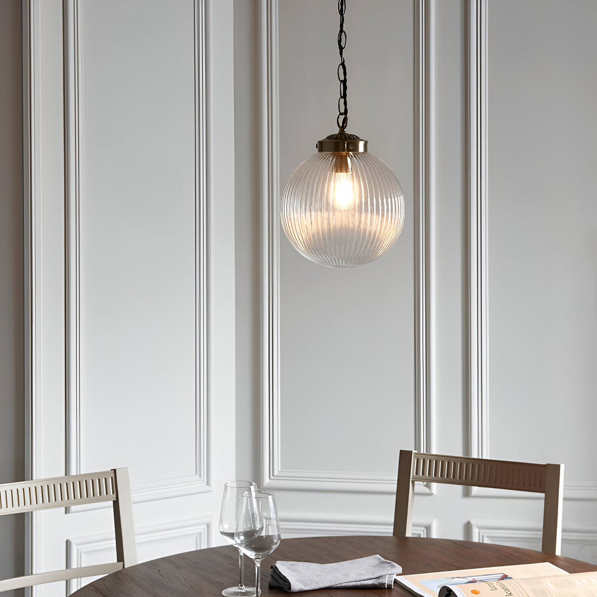 Endon Ashmere pendant with ribbed clear glass globe shade and antique brass chain, hung above a dining table with wine glasses against panelled walls.