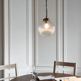 Endon Ashmere pendant with ribbed clear glass globe shade and antique brass chain, hung above a dining table with wine glasses against panelled walls.