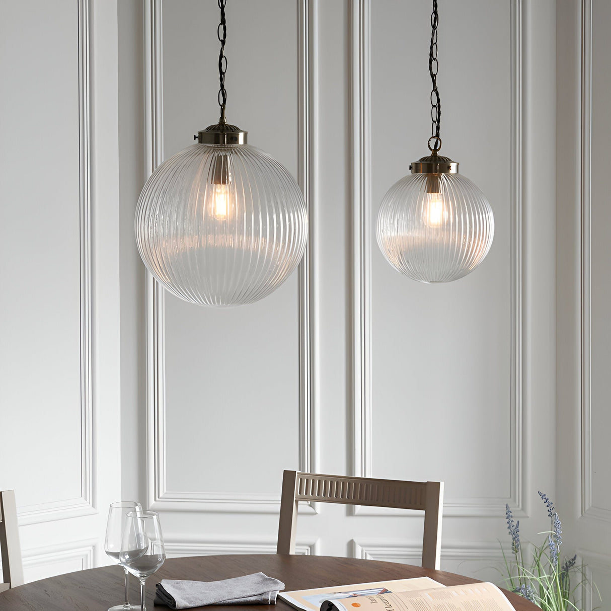 Endon Ashmere pendant lights in antique brass with ribbed clear glass globe shades on chains, hung over a dining table against panelled walls.