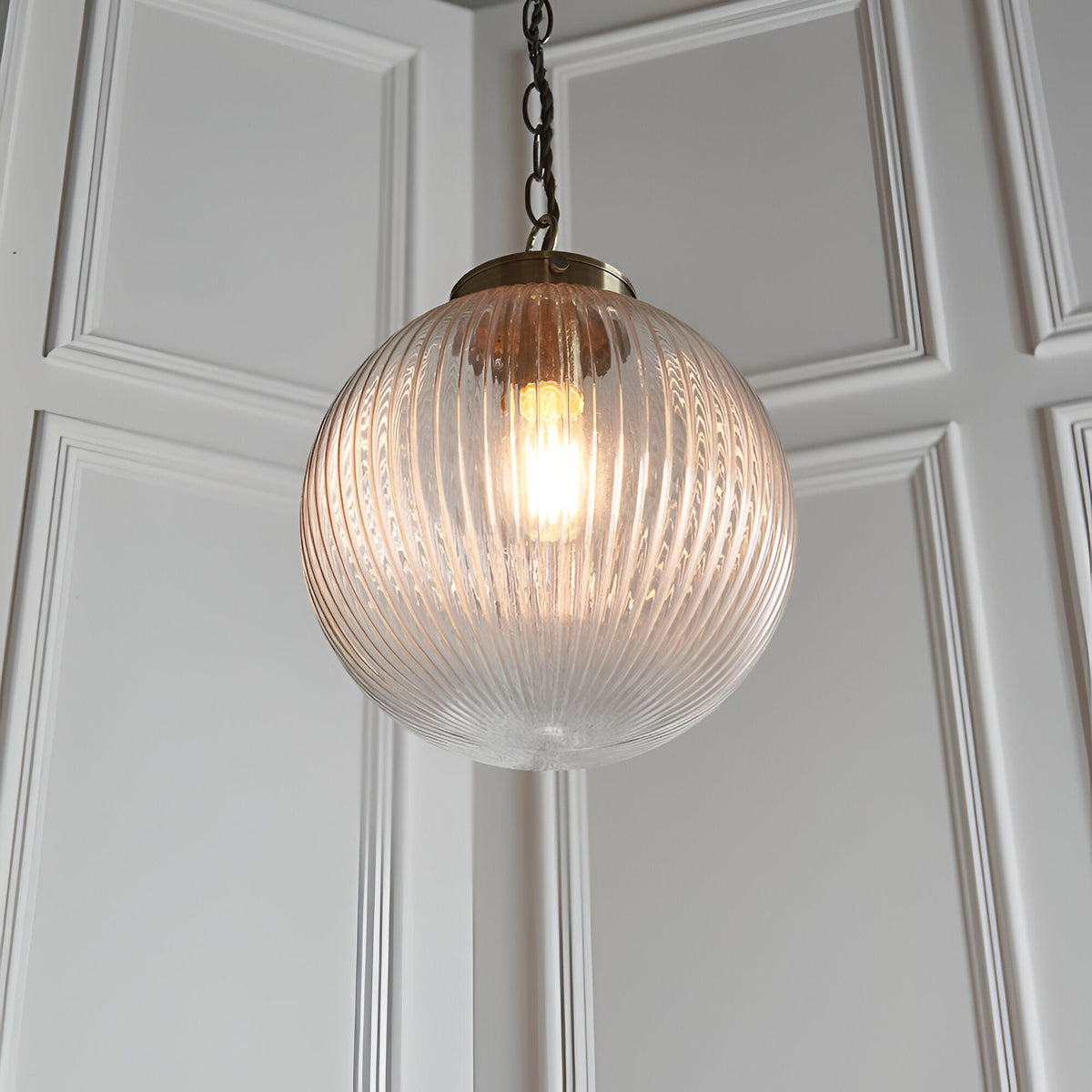 Endon Ashmere pendant light with ribbed round clear glass globe shade and antique brass chain suspension against white panelled wall.