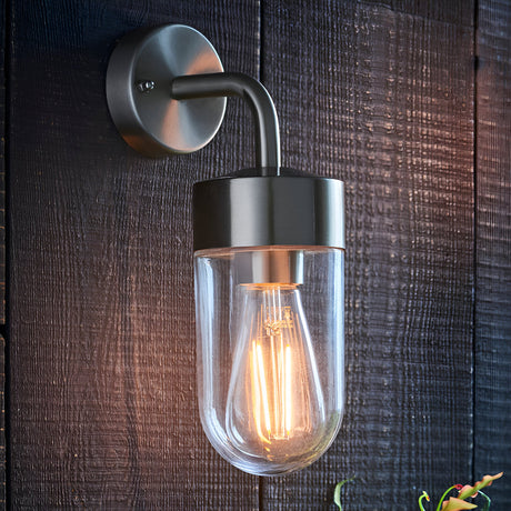 Endon Castfield outdoor wall light in brushed silver steel with a clear glass casing and exposed filament E27 bulb, mounted on a dark timber wall.