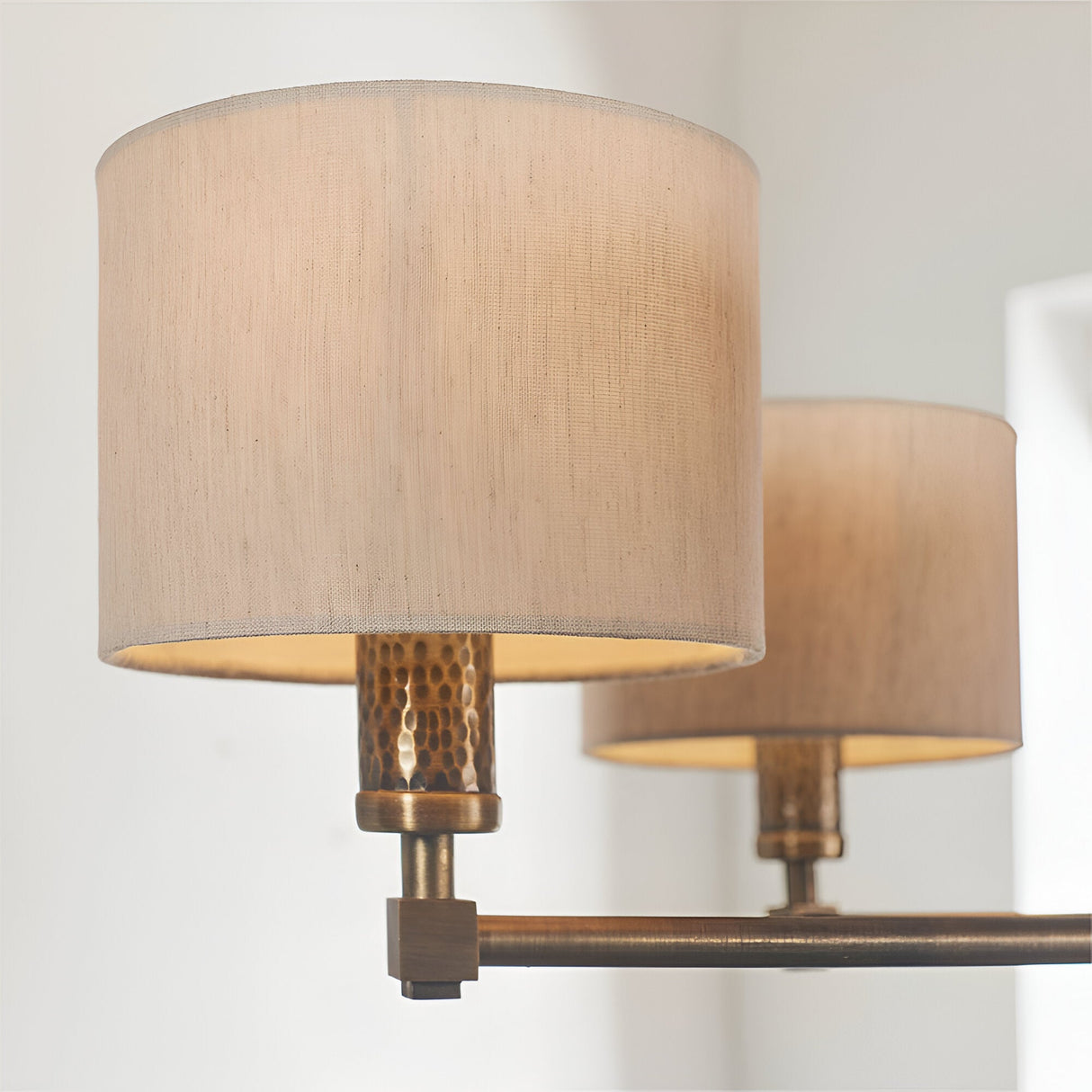 Close-up of Endon Brenwood pendant showing two natural linen drum shades on hammered bronze fittings with an aged bronze arm against a pale wall.