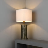 Endon Brenwood table lamp with hammered aged bronze cylindrical base and natural linen drum shade, lit on a grey console casting a warm glow.