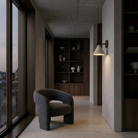A modern, minimalist hallway features large windows, a curved gray armchair, wooden shelves with ceramics, and the Trevino 1 Light Conical Travertine Wall Light in Walnut Brown & Light Beige. Neutral tones and city views at dusk complete the look.