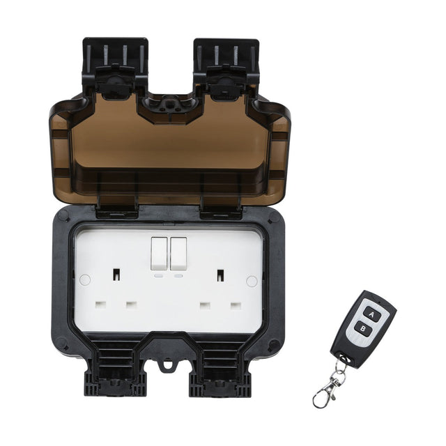 The Remote Controlled IP66 13A 2 Gang Outdoor Socket is displayed with its protective cover open, showcasing two UK-style sockets. Next to this sturdy configuration lies a compact remote control featuring buttons and a keychain attachment, perfect for effortlessly managing outdoor devices.