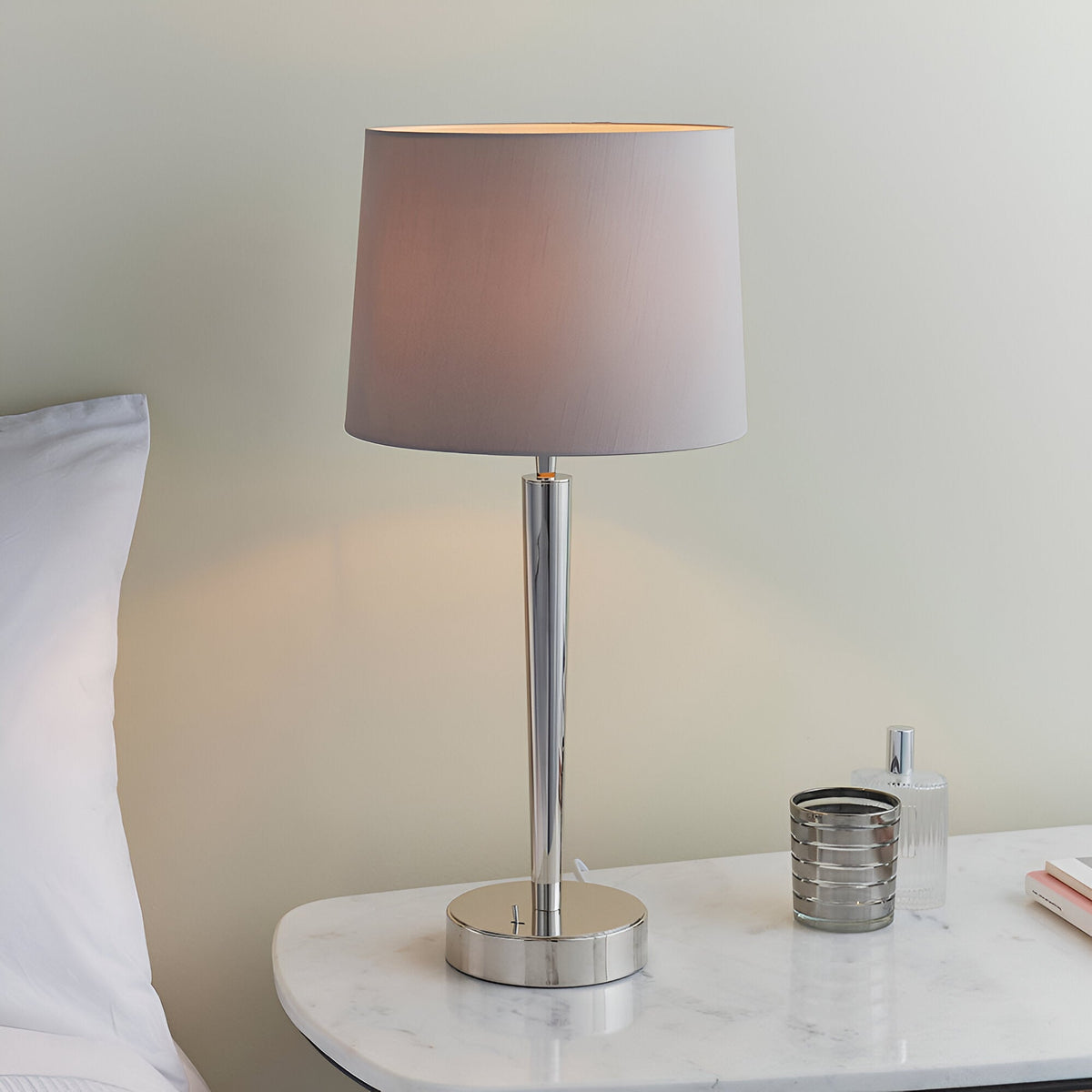 Endon Crandale USB table lamp with bright nickel stem and mink faux silk shade on a marble bedside table beside a bed and perfume bottle.