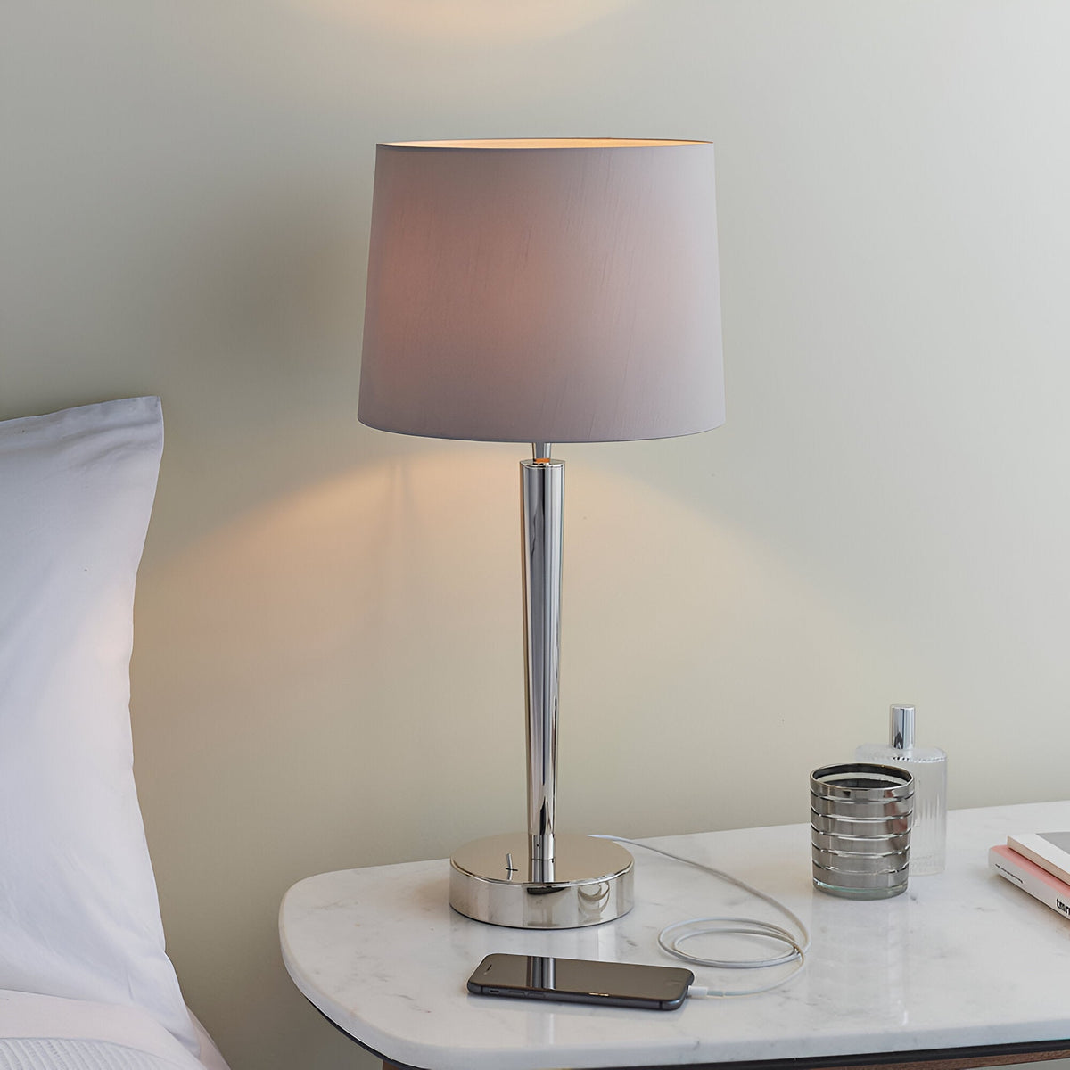 Endon Crandale USB table lamp in bright nickel with a mink faux silk shade, charging a phone on a marble bedside table.