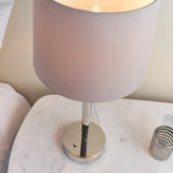 Endon Crandale USB table lamp with a bright nickel plated base, toggle switch and mink faux silk drum shade, shown lit on a marble bedside table.