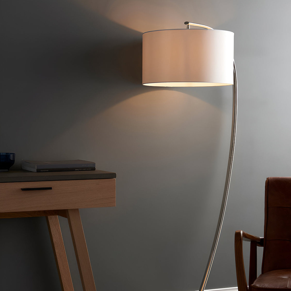 Brockfield Floor Lamp in polished nickel with an arched stem and large white faux silk drum shade, shown lit beside a wooden desk and leather chair.