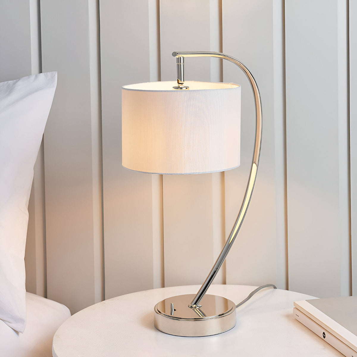Brockfield Table Lamp in nickel with an arched curved stem and white faux silk drum shade, shown on a bedside table.
