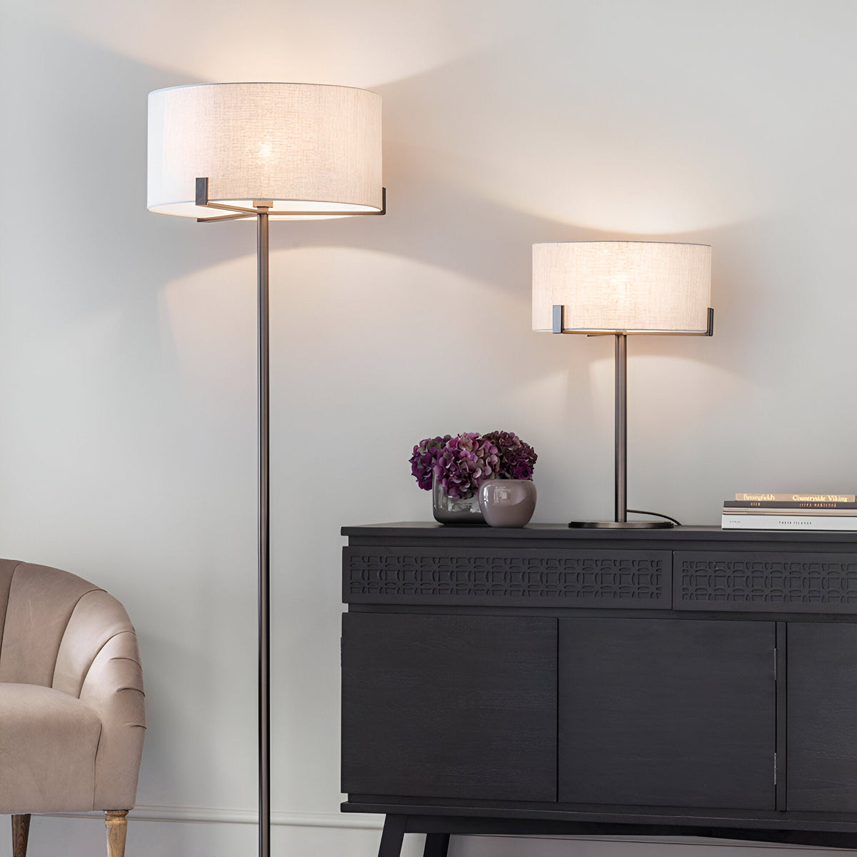 Brendon Table Lamp in bronze finish with a natural linen drum shade, shown lit on a dark sideboard alongside a matching floor lamp in a living room setting.