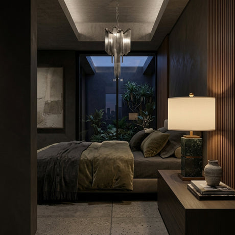 A modern, dimly lit bedroom showcases a double bed with dark bedding, the Veradex 1 Light Marble Block Table Lamp (Brass & Green) on the bedside table by a vase, an abstract painting, and a window revealing plants and a cityscape outside.