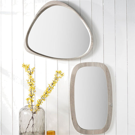 Grey oak wood veneer curved wall mirror, 67 x 58cm, with a rounded triangular frame, shown above a rectangular grey-framed mirror on white panelled wall with a vase of yellow flowers.