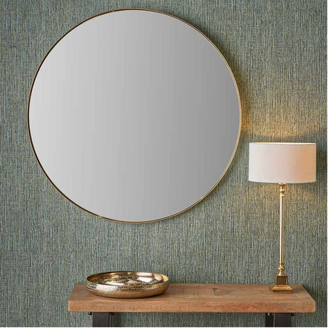 Round wall mirror with slim brushed gold frame, 100 x 100cm, shown above a wooden console table with a gold dish and brass table lamp against textured teal wallpaper.