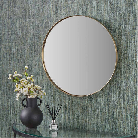 Round wall mirror with a slim brushed gold metal frame, 60 x 60cm, hung above a glass console table with a black vase and dried reeds against a textured teal wall.