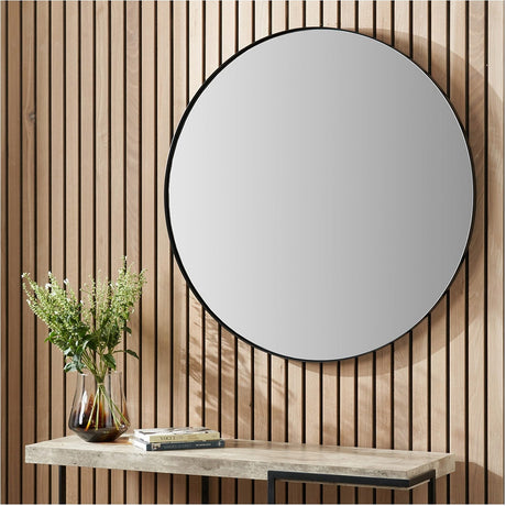Round wall mirror with slim black stainless steel frame, 100 x 100cm, hung on a wood-slatted wall above a console table with a vase and books.