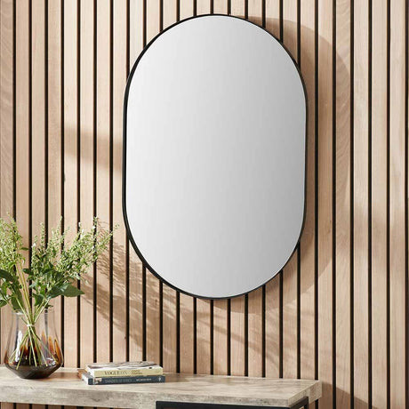 Oval wall mirror with a slim matt black stainless steel frame, 60 x 90cm, hung on a wood-slatted wall above a console table with plants and books.