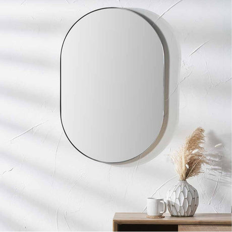 Oval wall mirror with a slim brushed silver stainless steel frame, 60 x 90cm, hung above a wooden sideboard with a vase and mug.
