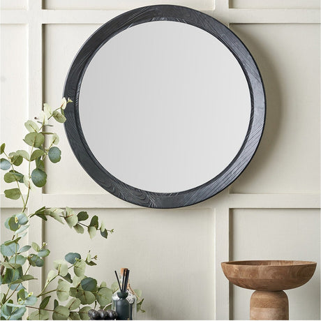 Elwood Black Fir Effect Round Wall Mirror, 65 x 65cm, with a textured black wood frame, shown on a cream panelled wall above eucalyptus foliage and a wooden bowl.