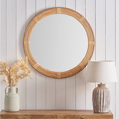 Round Rattan Frame Wall Mirror, 80 x 80cm, in natural woven finish, hung on a white panelled wall above a wooden sideboard with a vase and lamp.
