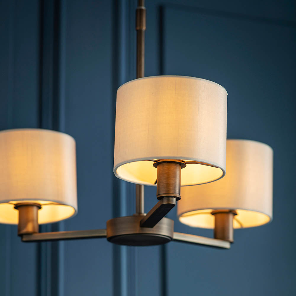 Endon Bexley 3 light pendant in antique bronze with three marble-effect faux silk drum shades on curved arms, shown lit against a deep blue panelled wall.
