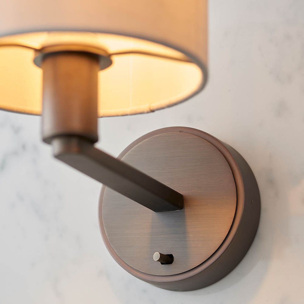 Endon Bexley wall light close-up showing the bronze brushed arm, circular backplate with on/off toggle switch, and fabric shade against a marble wall.