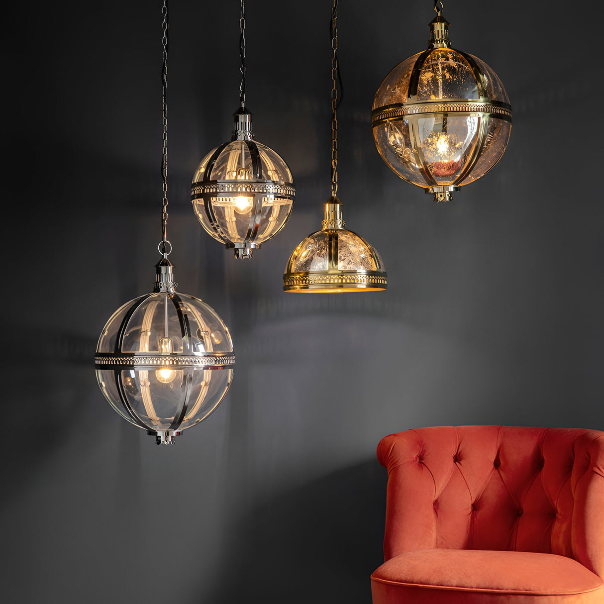 Endon Danecombe pendant lights in brass and nickel with clear glass globes and a dome shade, grouped above a red tufted armchair against a dark wall.